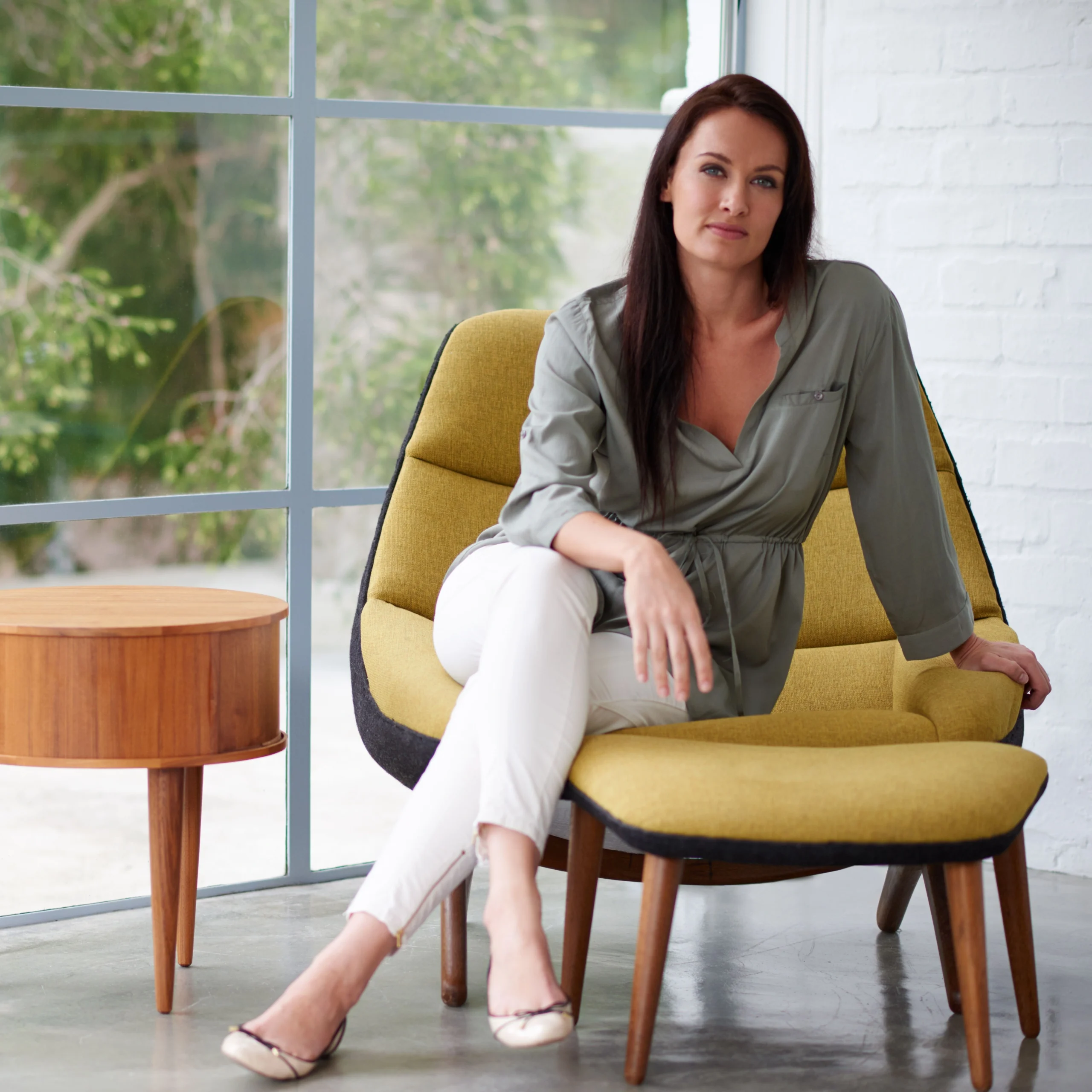 A woman in a stylish green blouse and white pants sits confidently on a modern yellow chair near a large window, exuding relaxation and elegance.