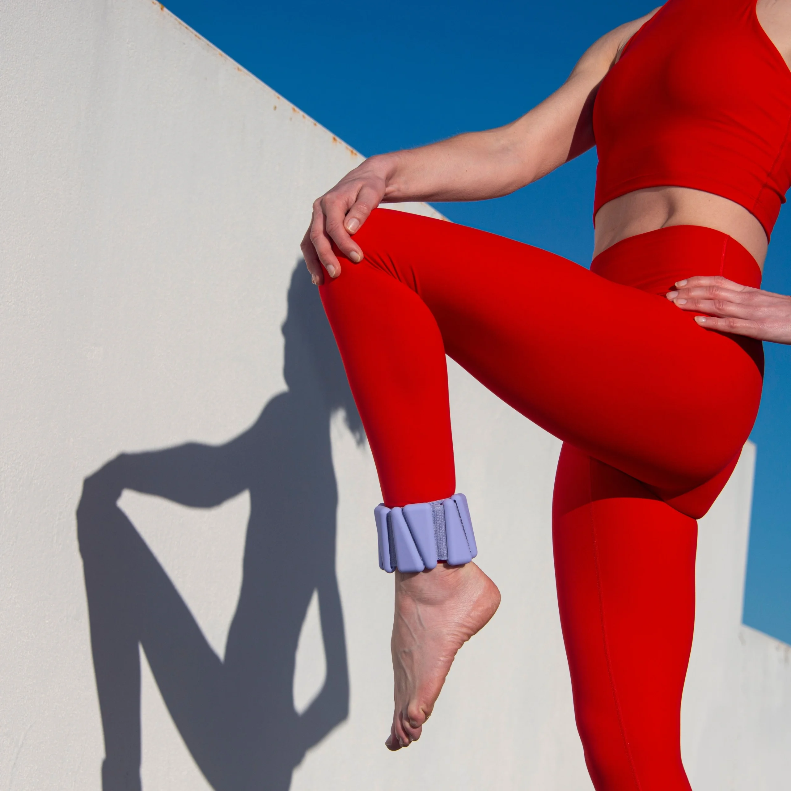A woman in a red athletic outfit performs a balancing exercise with ankle weights against a bright blue sky, symbolizing strength and fitness.