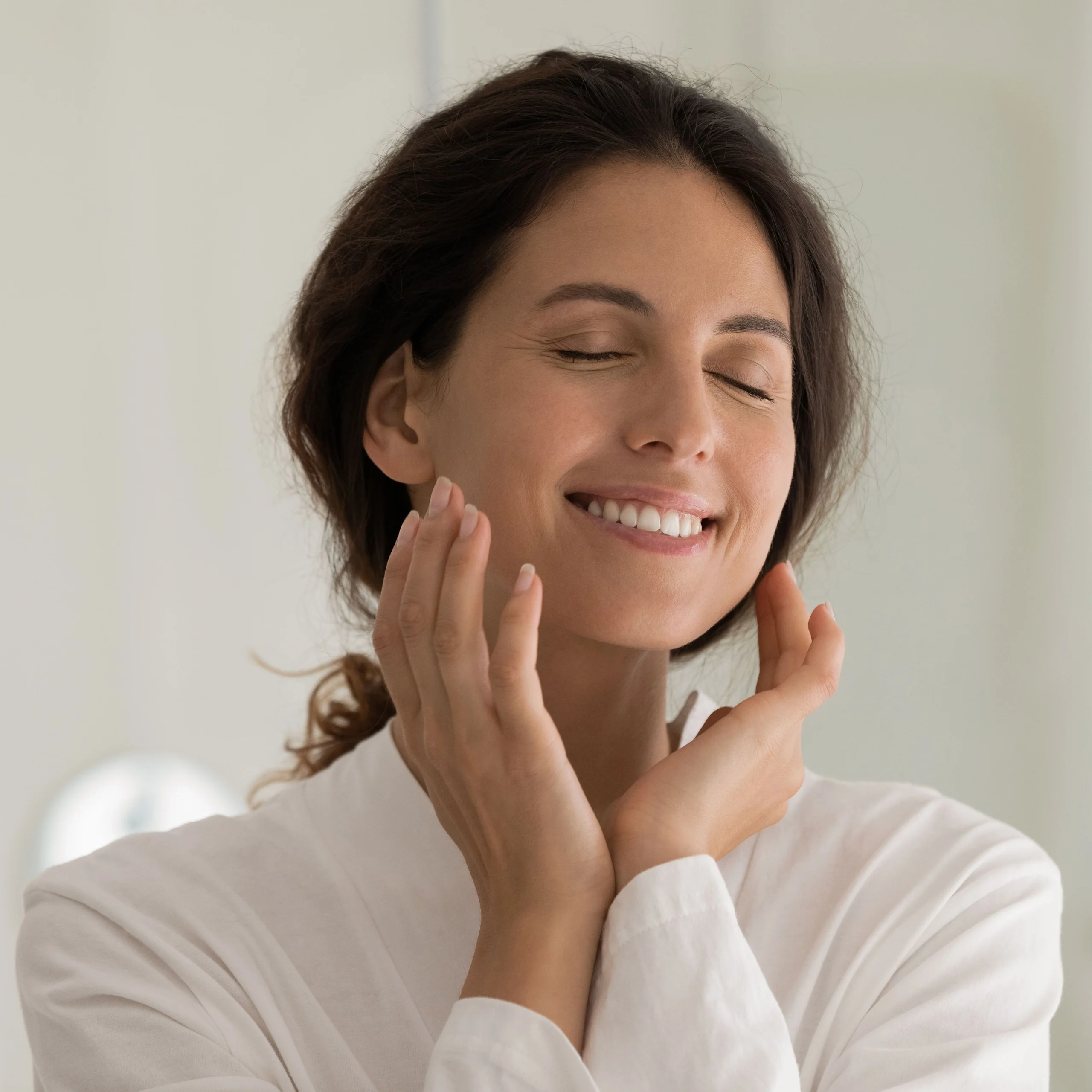 A woman in a white robe gently touches her face with a serene smile and closed eyes, appearing to enjoy a skincare or facial treatment.