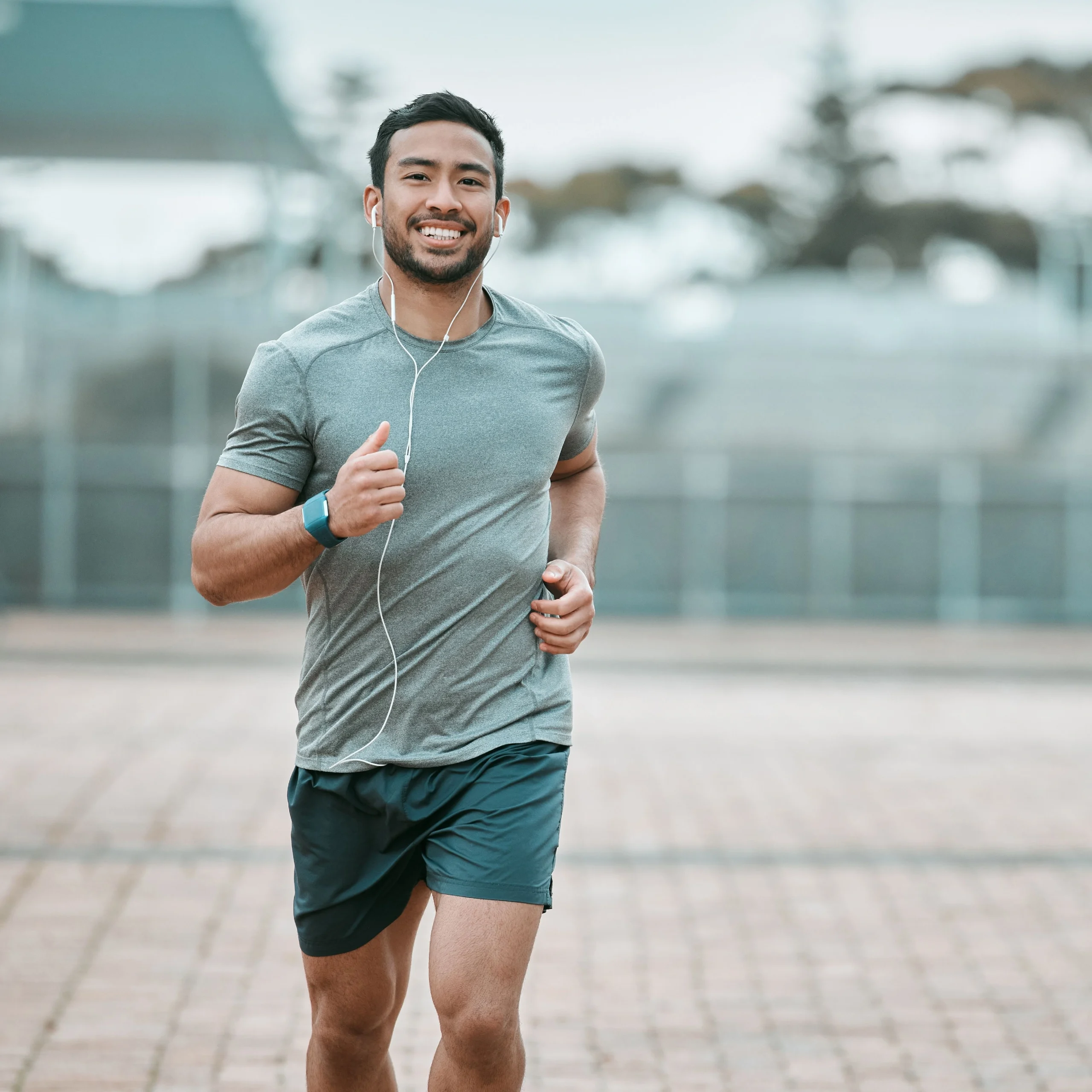 A smiling man in athletic wear jogs outdoors while listening to music through earphones, exuding energy and fitness.